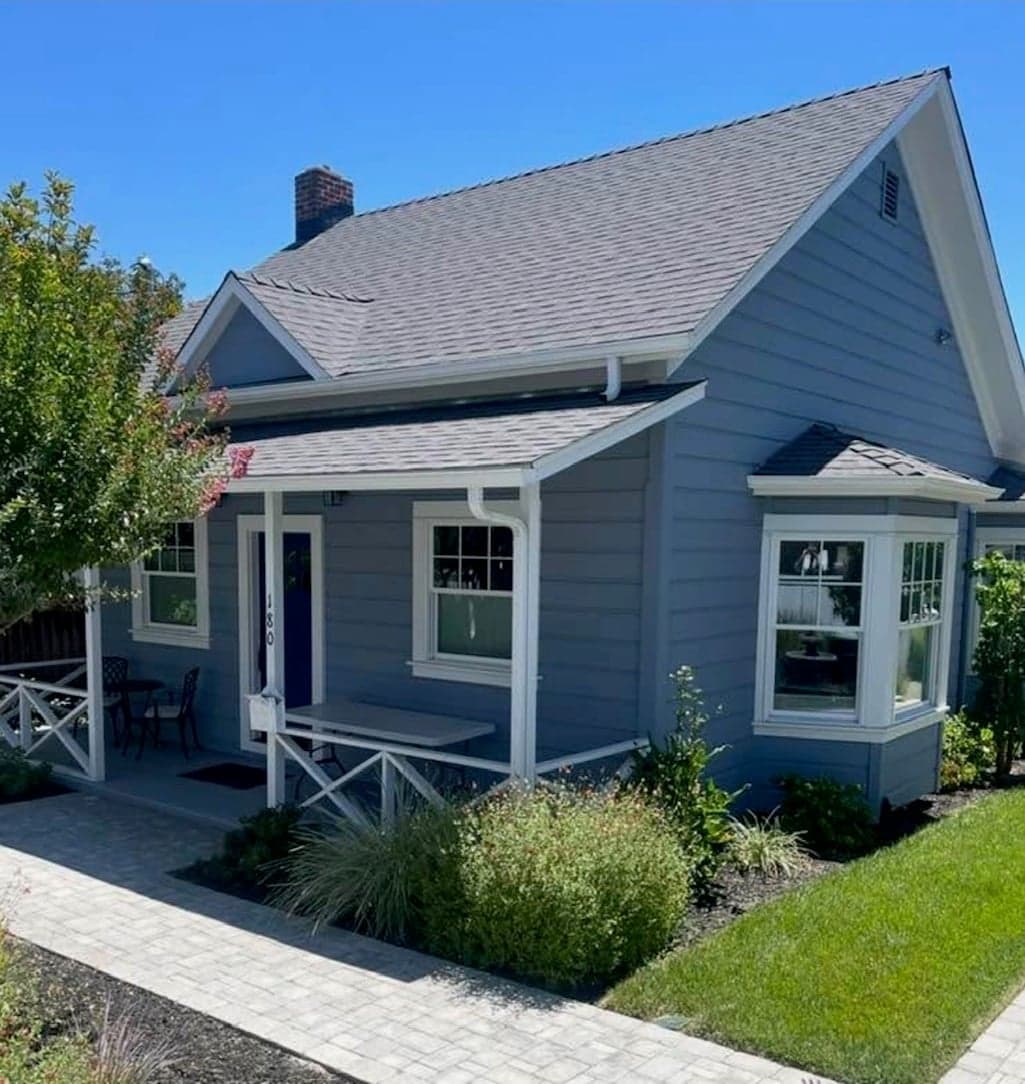 The restored single-storey house in blue-grey siding with white trim, a steep gable, a bay window, and a small covered front porch behind new landscaping and a paver walk