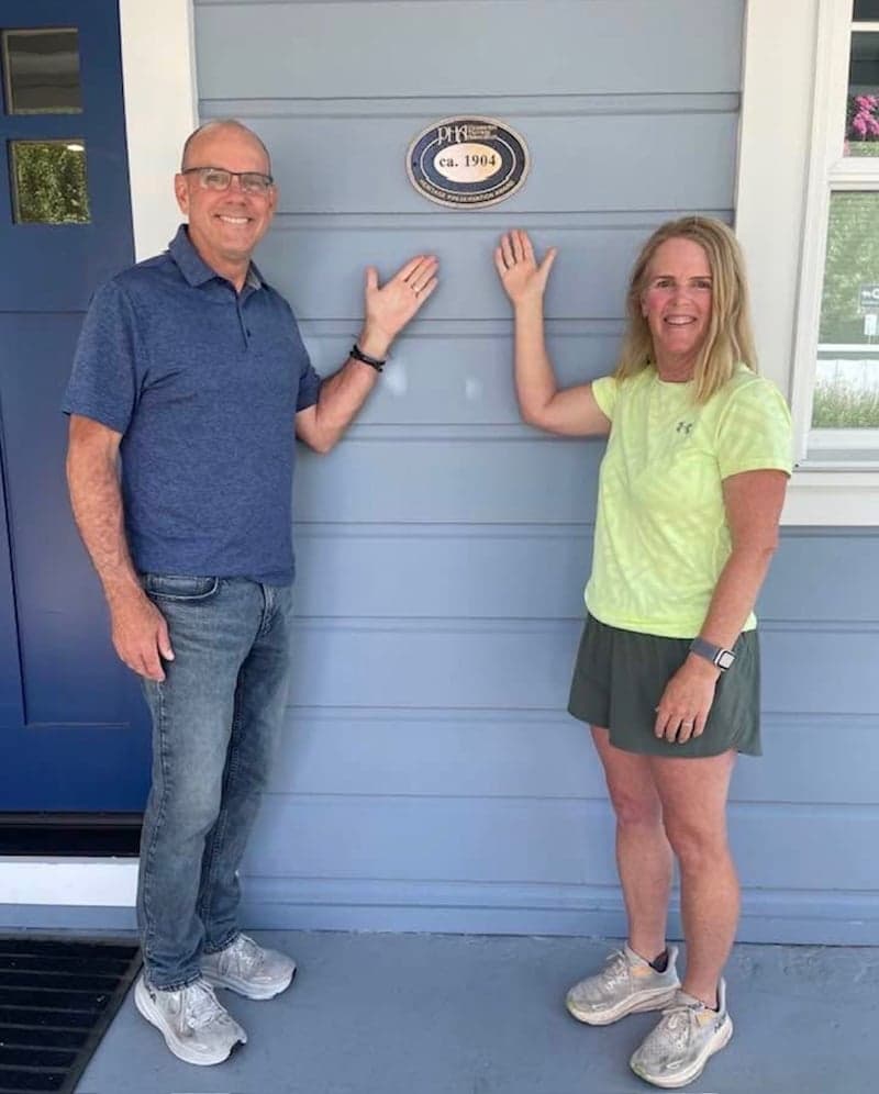 Shawn and Dan Copenhagen standing on either side of the association's ca. 1904 Heritage Preservation Award plaque, mounted on the house's blue-grey siding