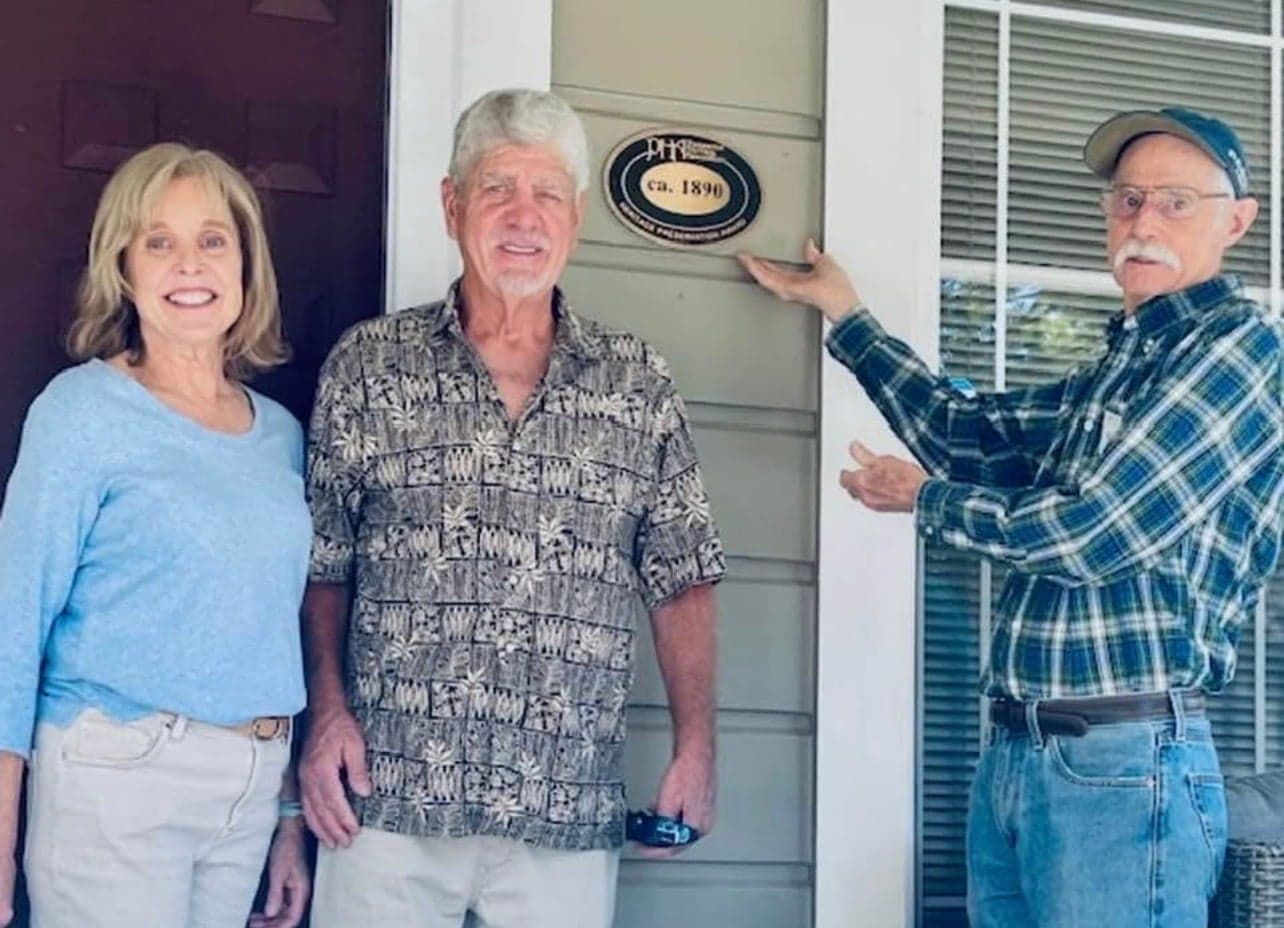 George and Jeanie Emmett on their porch beside the association's ca. 1890 Heritage Preservation Award plaque, mounted on grey siding, while a PHA member gestures toward it