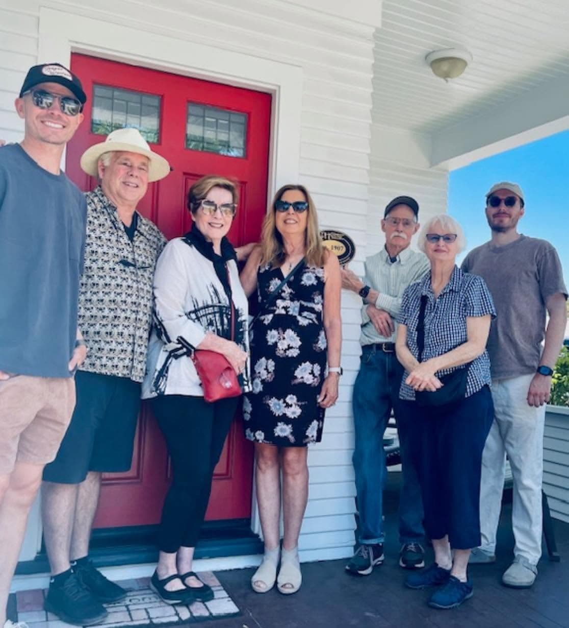 Seven people standing together on the porch in front of the red double door, with the ca. 1907 plaque on the siding behind them