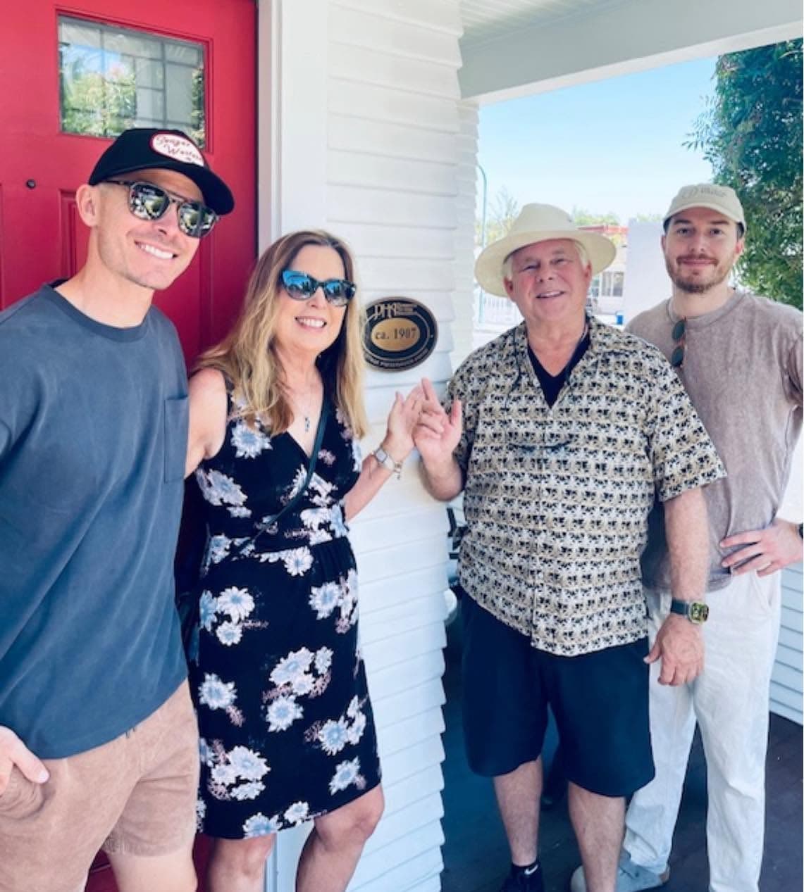 Four members of the Knuppe family on the front porch beside the red door, gathered around the association's ca. 1907 Heritage Preservation Award plaque on the white siding