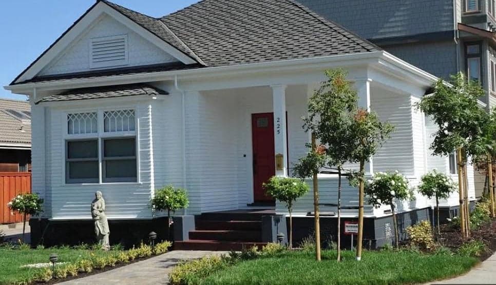 The restored single-storey cottage in white horizontal siding with a red front door, a gabled front bay with diamond-paned upper windows, a covered corner porch, and young trees and new lawn along a paver walk