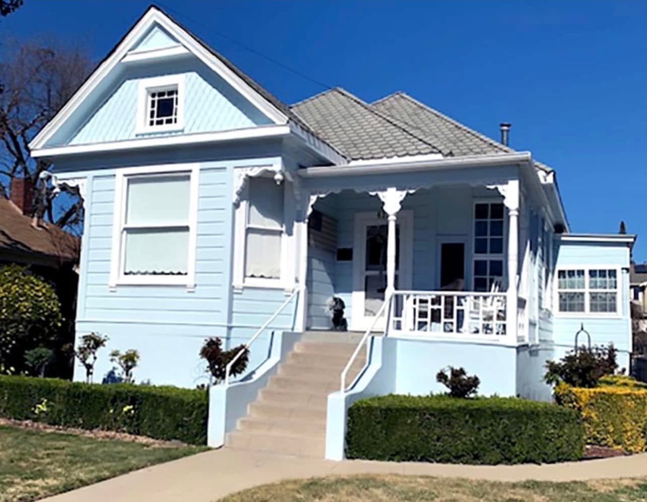 A pale blue single-storey cottage with a steep front gable, white scalloped trim, and a corner porch reached by a tall flight of concrete steps