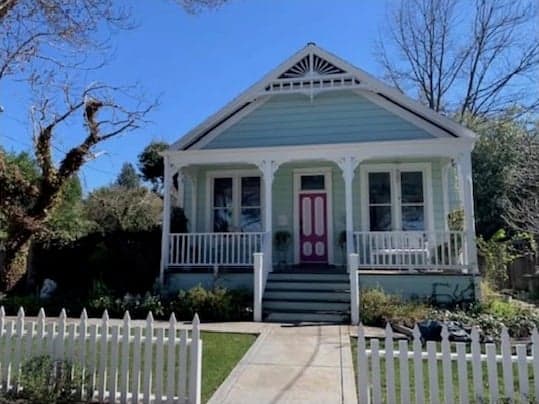 A single-storey end-gable cottage in pale blue with white trim, a sunburst in the gable peak, a full-width porch with turned posts, a pink front door, and a white picket fence