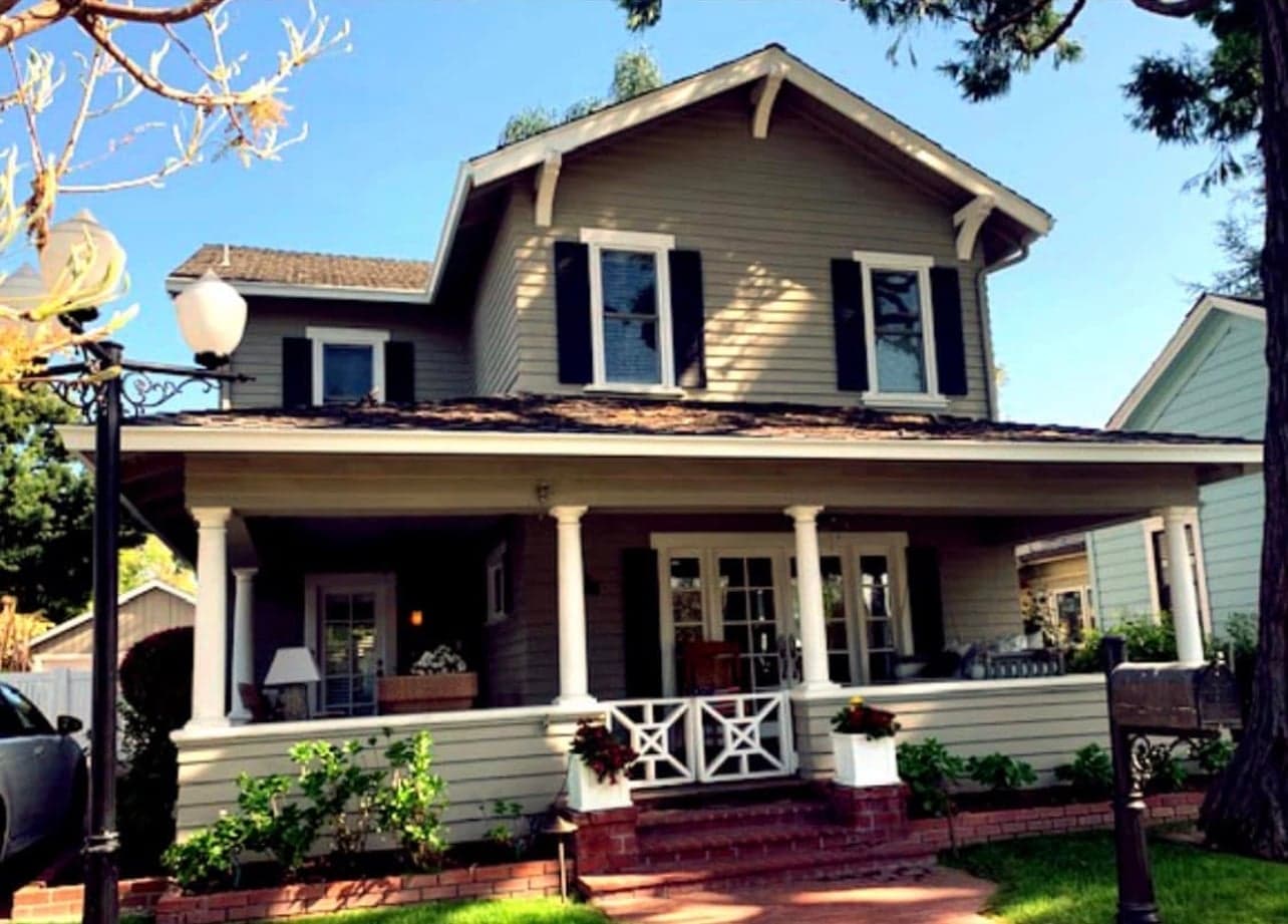 A two-storey grey clapboard house with black shutters, white bracketed gables, and a deep front porch on white columns behind a brick-edged garden