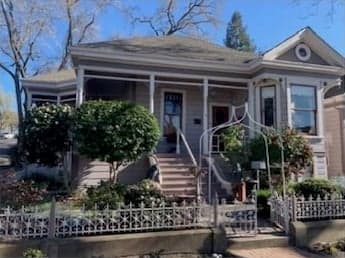 A grey Queen Anne cottage with white trim, a gabled bay with a round vent, and a front porch reached by steps through a white garden arch and ornamental iron fence