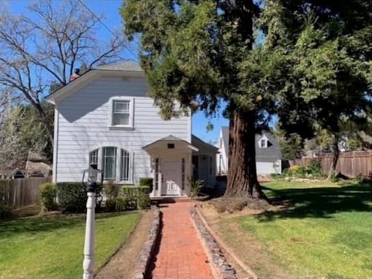 A white two-storey end-gable house with a small gabled entry porch, reached by a long brick path past a lamp post, beside a very large redwood tree