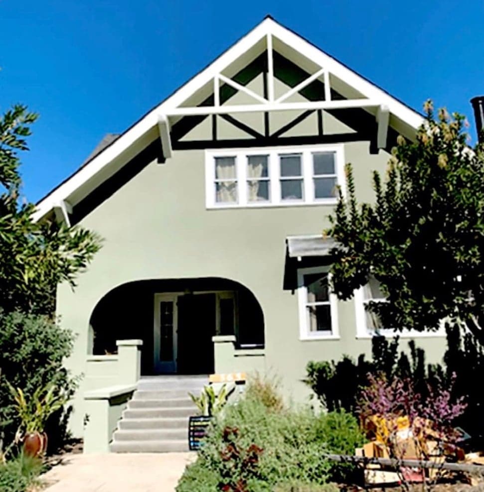 A sage-green stucco house with a steep front gable of white and dark timber bracing, a bank of four upstairs windows, and an arched entry porch above concrete steps