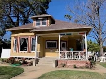 A yellow hip-roofed cottage with a small dormer, a red-trimmed bay window, and a corner porch with white railings above a brick base